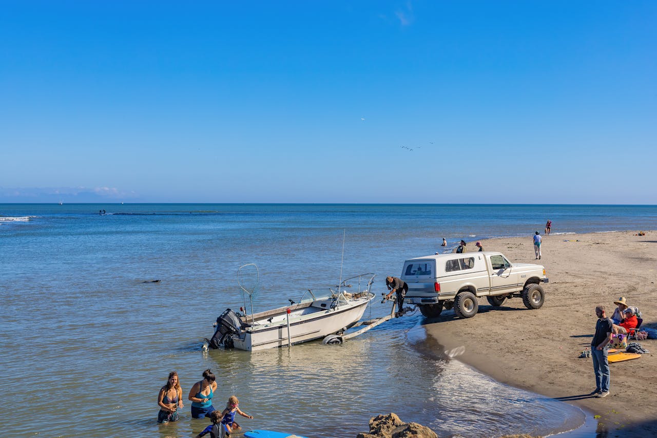 A truck towing a boat on a sunny seaside with people enjoying the beach.
